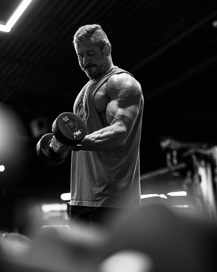 Black and white photo of a muscular man lifting dumbbells in a gym setting.